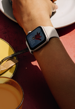 Lidská paže sahající po snídani, Apple Watch SE 3 na zápěstí, pouzdro z hvězdně bílého hliníku, stále zapnutý displej, na ciferníku lidský portrét, hvězdně bílý sportovní řemínek