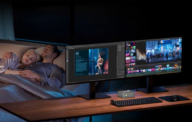 Mini PC na stole v noci vedle dvou monitorů; spící pár v pozadí naznačuje tichý provoz zařízení.