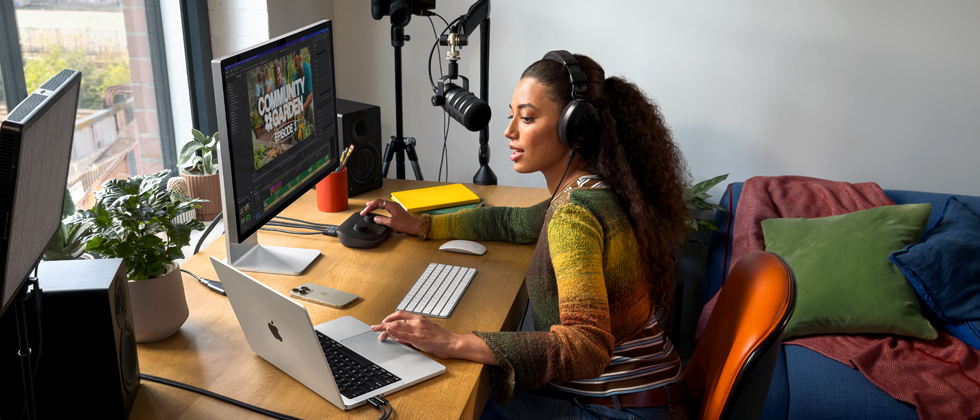 Žena doma pracuje u svého stolu na MacBooku Pro připojeném ke dvěma externím monitorům, má nasazená sluchátka a mluví do mikrofonu